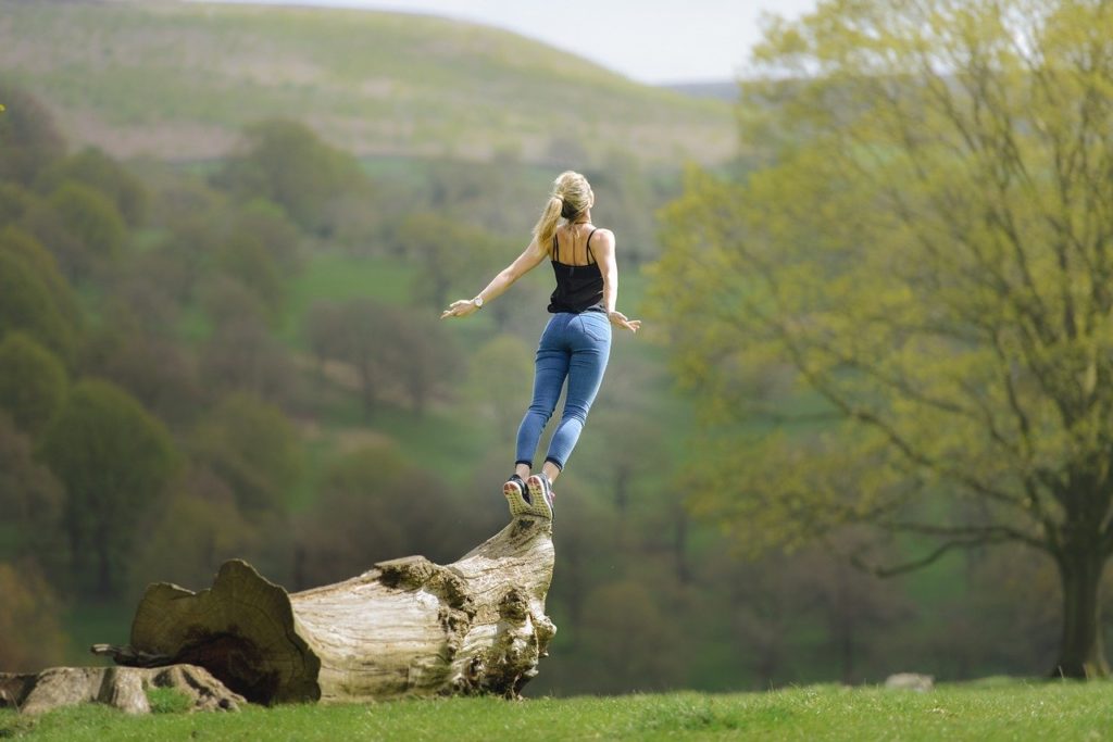 femme sautant sur une bûche dans un paysage montagneux verdoyant, bien-être actif en nature