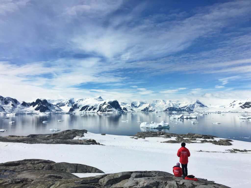 deux voyageurs en rouge face aux icebergs et montagnes enneigées de l'antarctique