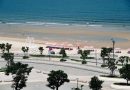 A sunny beach with pink umbrellas, clear blue ocean, and a promenade in the foreground.