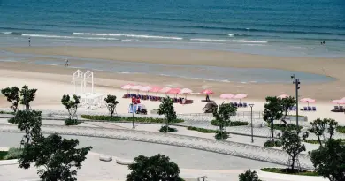 A sunny beach with pink umbrellas, clear blue ocean, and a promenade in the foreground.